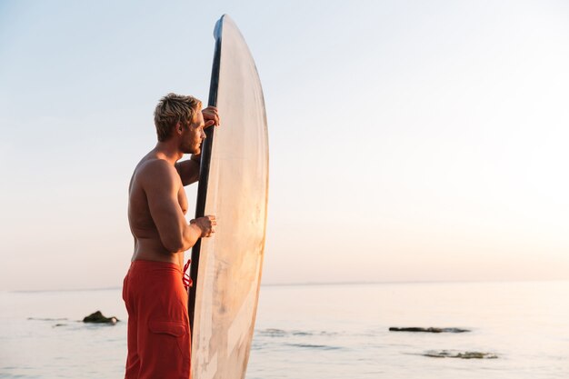 Zdjęcie blond pół koszuli wysportowany surfer trzymający swoją deskę surfingową nad oceanem o wschodzie słońca