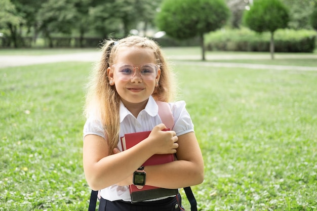 blond uśmiechnięta uczennica w szkolnym mundurku trzymająca notatnik z różowym plecakiem z powrotem do szkoły