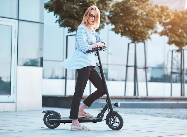 Blond Zamyślona Kobieta W Okularach Prowadzi Swój Nowy Skuter Elektryczny Na Ulicy W Jasny, Słoneczny Dzień.