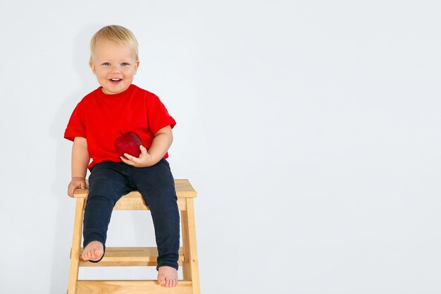 Blondwłose dziecko w czerwonej koszulce uśmiecha się, trzymając w dłoni jabłko, siedząc na krześle