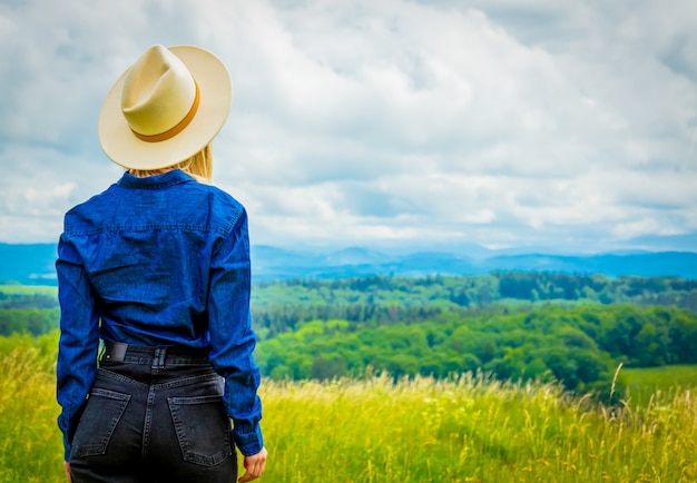 Blondynka cowgirl w kapeluszu na łące z górami w tyle
