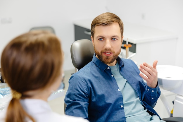 Zdjęcie blondynka dentysta w gabinecie dentystycznym rozmawia z pacjentem płci męskiej i przygotowuje się do leczenia przystojny brodaty mężczyzna w fotelu dentystycznym patrząc na swojego lekarza z uśmiechem