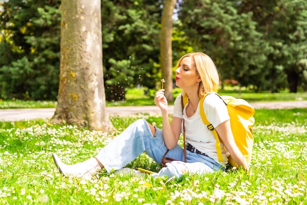 Blondynka dmuchająca na roślinę mniszka lekarskiego w parku w mieście, w kapeluszu i okularach przeciwsłonecznych, siedząca wiosną na trawie obok stokrotek