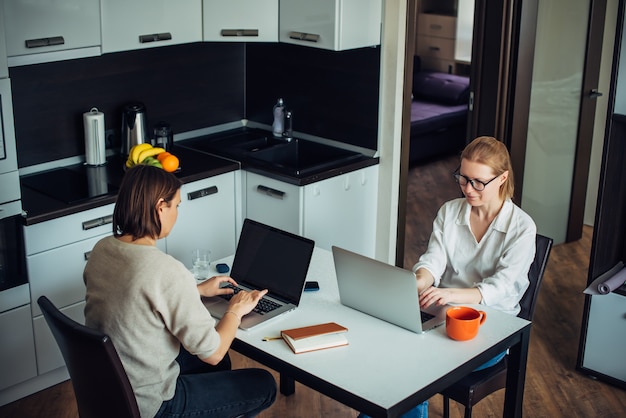 Blondynka i brunetka pracują na laptopach, siedząc naprzeciw siebie przy stole w kuchni. Coworking w przytulnym domowym wnętrzu.