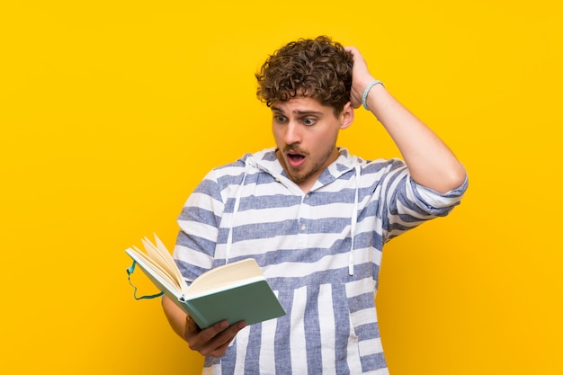 Blondynka mężczyzna nad kolor żółty ścianą zaskakującą podczas gdy cieszący się czytający książkę