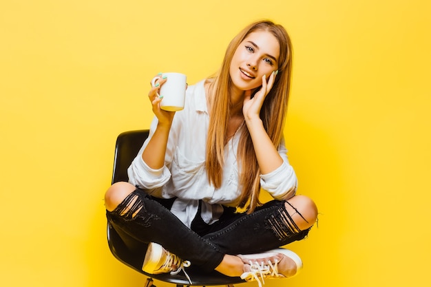 Blondynka młoda kobieta na izolowanej ścianie żółty, ubrany w odzież biznesową, trzymając filiżankę gorącej kawy i smilimg.