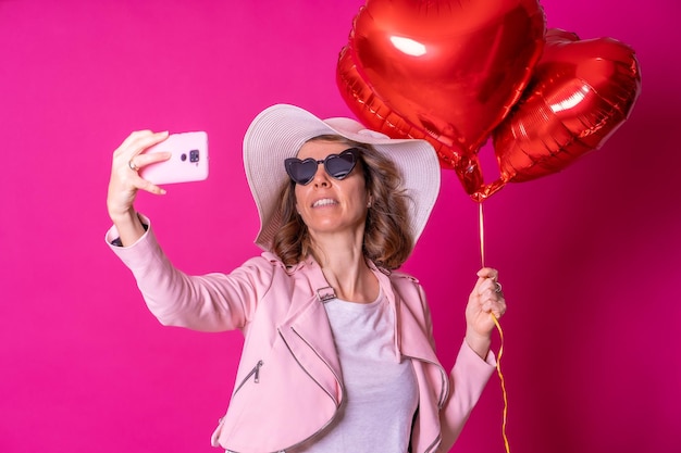 Blondynka rasy kaukaskiej bawi się z białym kapeluszem i okularami przeciwsłonecznymi w klubie nocnym z balonami w kształcie serca, biorącymi selfie z różowym tłem telefonu komórkowego