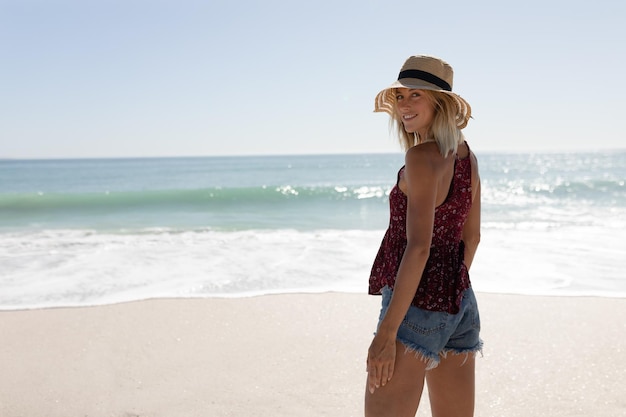 Blondynka rasy kaukaskiej ubrana w słomkowy kapelusz i dżinsowe szorty, ciesząca się wolnym czasem spacerując nad morzem na idyllicznej słonecznej plaży, obracając się i uśmiechając do kamery. Relaksujące wakacje na tropikalnej plaży