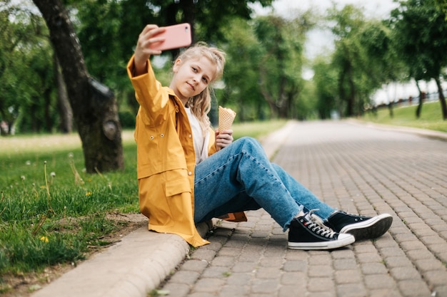 Zdjęcie blondynka siedząca na drodze z telefonem i lodem robi zabawne selfie dziecka na świeżym powietrzu