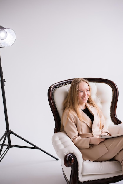 Zdjęcie blondynka siedząca na krześle w białym studiu fotograficznym, ubrana w formalny beż.
