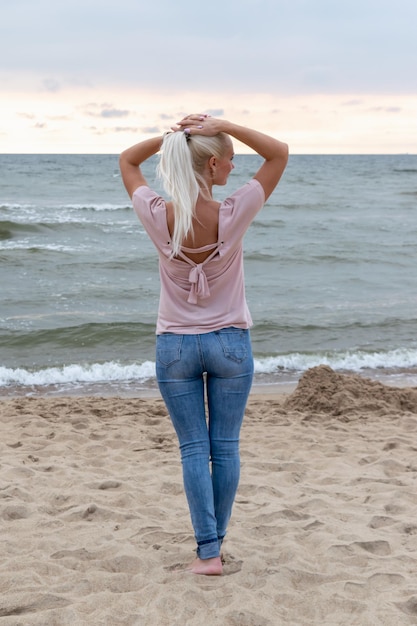 Blondynka stoi na plaży nad morzem z rękami za głową i patrzy na zachód słońca