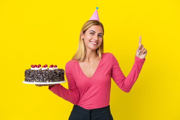 Blondynka Urugwajska trzyma tort urodzinowy na białym tle na żółtym tle pokazując i podnosząc palec na znak najlepszych