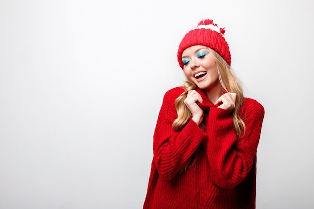 Blondynka uśmiecha się w czerwonym swetrze i czapce z jasnym zimowym makijażem na szarym tle