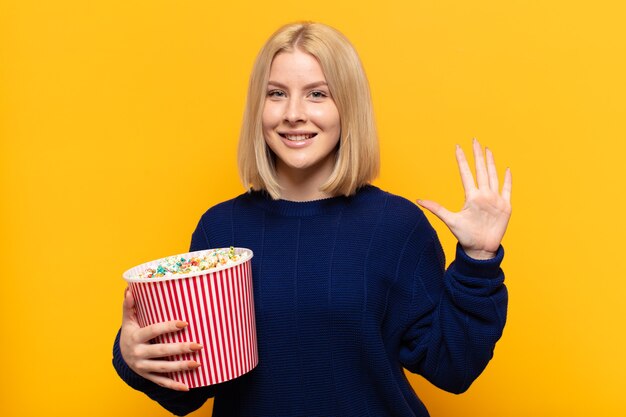 Blondynka uśmiechnięta i wyglądająca przyjaźnie, pokazująca numer pięć lub piąty z ręką do przodu, odliczający