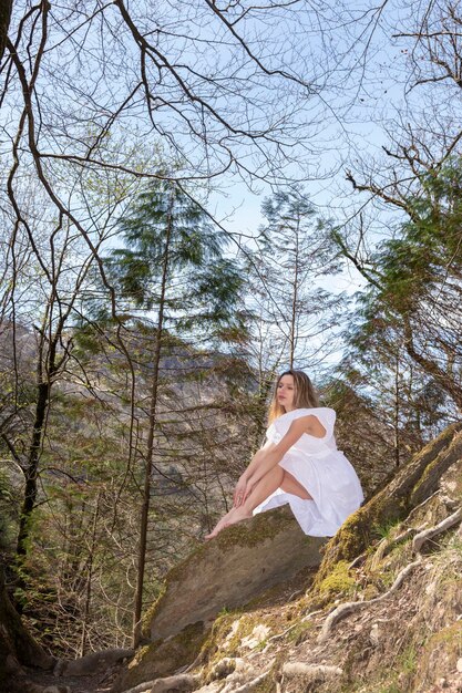 Zdjęcie blondynka w białej sukience siedząca na skale w lesie.