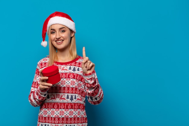 Blondynka w czapce Świętego Mikołaja uśmiechnięta i wyglądająca przyjaźnie, pokazująca numer jeden lub pierwszy z ręką do przodu, odliczająca