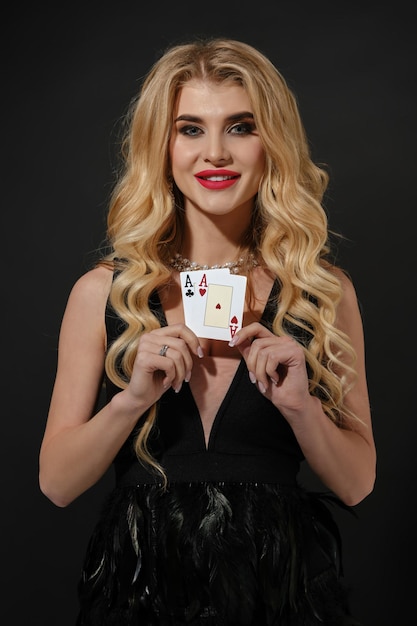 Blondynka w czarnej stylowej sukience i naszyjniku uśmiecha się pokazując dwie karty do gry pozujące na b...