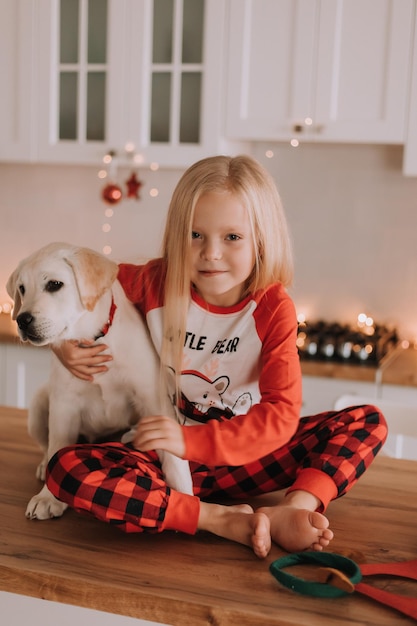 Blondynka w czerwonej świątecznej piżamie siedzi w ozdobionej girlandą kuchni z białym psem. zimowe weekendy i święta w ciepłym rodzinnym gronie. dziecko i zwierzę. miejsce na tekst. Zdjęcie wysokiej jakości