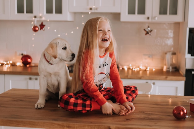 Blondynka w czerwonej świątecznej piżamie siedzi w ozdobionej girlandą kuchni z białym psem. zimowe weekendy i święta w ciepłym rodzinnym gronie. dziecko i zwierzę. miejsce na tekst. Zdjęcie wysokiej jakości