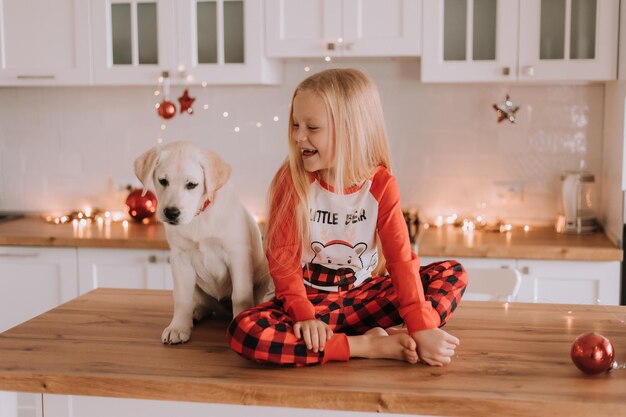 Blondynka w czerwonej świątecznej piżamie siedzi w ozdobionej girlandą kuchni z białym psem. zimowe weekendy i święta w ciepłym rodzinnym gronie. dziecko i zwierzę. miejsce na tekst. Zdjęcie wysokiej jakości