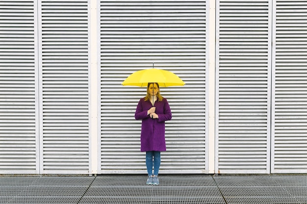 Blondynka w fioletowym płaszczu i żółtym parasolem pozuje przed białą ścianą o geometrycznej strukturze.