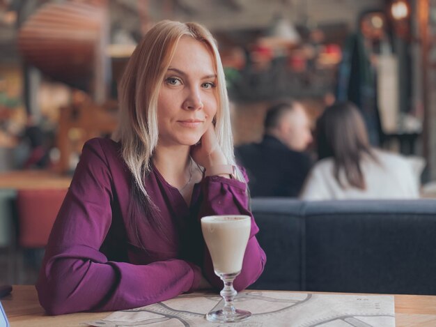 Blondynka w liliowym swetrze siedzi na sofie w przytulnej restauracji, jest zrelaksowana i uśmiechnięta i pije kawę na swoim stole.
