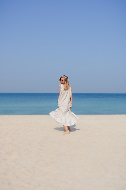 Blondynka w lnianej sukience maxi z rozwianymi włosami, skacząca i tańcząca na piaszczystej plaży