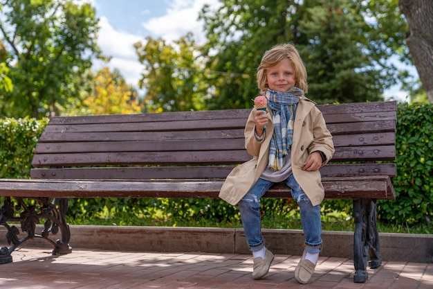 Blondynka w płaszczu i szaliku je lody siedząc w parku na drewnianej ławce