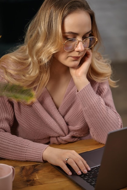 Blondynka w różowym swetrze i okularach. Podpiera twarz dłonią, pracując na laptopie. Siedząc przy drewnianym stole z różową filiżanką. Blogger. Wnętrze z białym murem. Ścieśniać