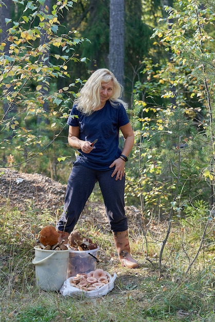 Blondynka z nożem w dłoni i rozpuszczonymi włosami stoi w lesie obok zebranych grzybów