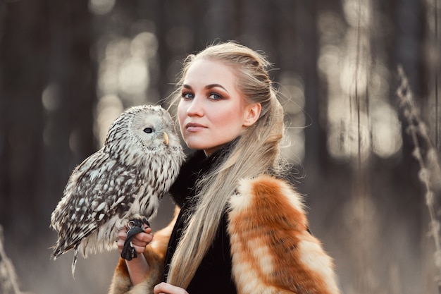 Blondynki kobieta z sową w jej rękach chodzi w lesie jesienią i wiosną. Długie włosy kobieta, romantyczny portret z sową. Fotografia mody, piękny makijaż