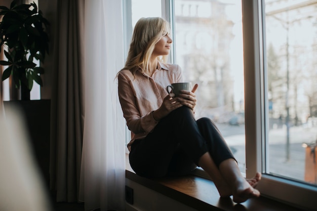 Blondynki młoda kobieta relaksuje kawę i pije okno