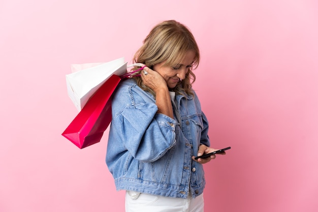 Blondynki w średnim wieku na odizolowanych różowej ścianie trzymając torby na zakupy i pisząc wiadomość swoim telefonem komórkowym do przyjaciela