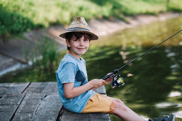 Boczny widok szczęśliwa chłopiec jest ubranym kapeluszowego połów na jeziorze