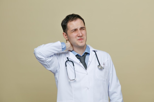 Bolący młody mężczyzna lekarz ubrany w szatę medyczną i stetoskop wokół szyi trzymający rękę za szyją z zamkniętymi oczami odizolowanymi na oliwkowym tle