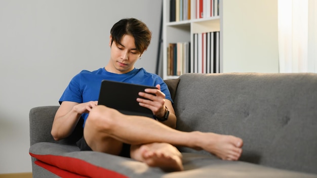 Boso młody azjatycki mężczyzna w swobodnym stroju siedzi na kanapie i przegląda internet na cyfrowym tablecie
