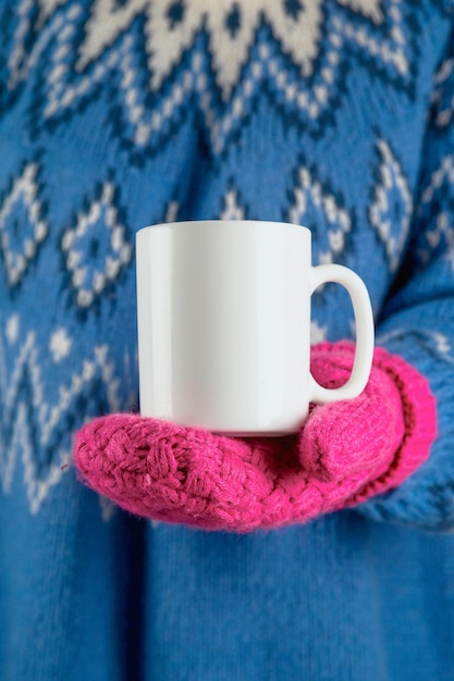 Boże Narodzenie biały kubek makieta w dłoni na kobiecie noszącej różowe rękawiczki i niebieski sweter.
