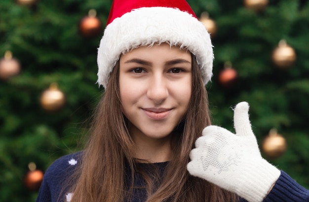 Boże Narodzenie jak kciuk w górę. Ścieśniać Portret kobiety w kapeluszu Santa Claus z emocjami. Na tle choinki.