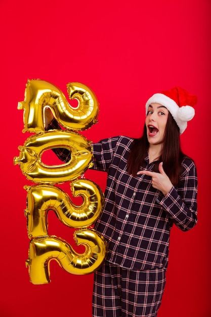 Boże Narodzenie, koncepcja wesołych wakacji - młoda kobieta w piżamie i czapce Świętego Mikołaja trzyma cyfry 2022