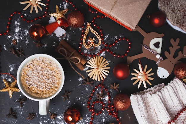 Boże Narodzenie płaskie ułożenie gorącej kawy lub kakao na czarnym drewnianym tle z ręcznie robionymi zabawkami świątecznymi, czerwonymi kulkami, koralikami, goździkami i cynamonem. Zbliżenie, widok z góry