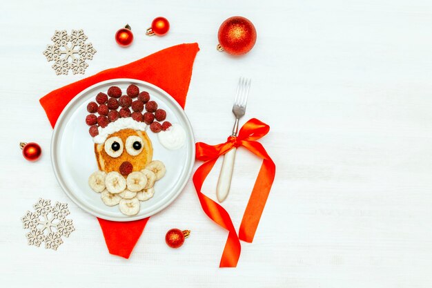 Boże Narodzenie Santa Claus twarz w kształcie naleśnika z jagodami słodkich świeżych malin i bananem na talerzu na białym tle drewnianych dla dzieci dzieci śniadanie. Boże Narodzenie deser żywności z dekoracjami na nowy rok.