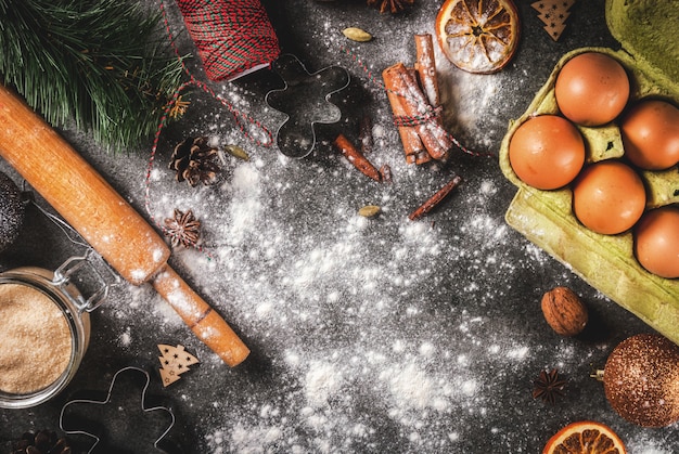 Boże Narodzenie, świąteczne gotowanie. Składniki, przyprawy, suszone pomarańcze i formy do pieczenia, ozdoby świąteczne (kule, gałąź jodły, szyszki), na czarnym kamiennym stole, widok z góry
