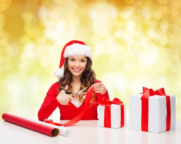 Boże Narodzenie, święta, Celebracja, Dekoracja I Koncepcja Ludzi - Uśmiechnięta Kobieta W Kapeluszu Pomocnika świętego Mikołaja Z Nożyczkami Pakująca Pudełko Na żółtym Tle światła
