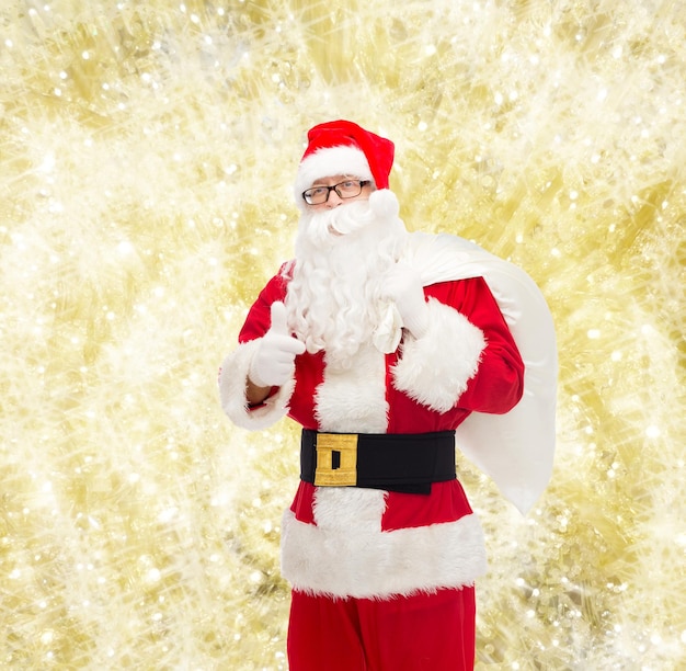 Boże Narodzenie, święta, gest i koncepcja ludzi - mężczyzna w stroju świętego mikołaja z torbą pokazującą kciuki w górę na żółtym tle światła