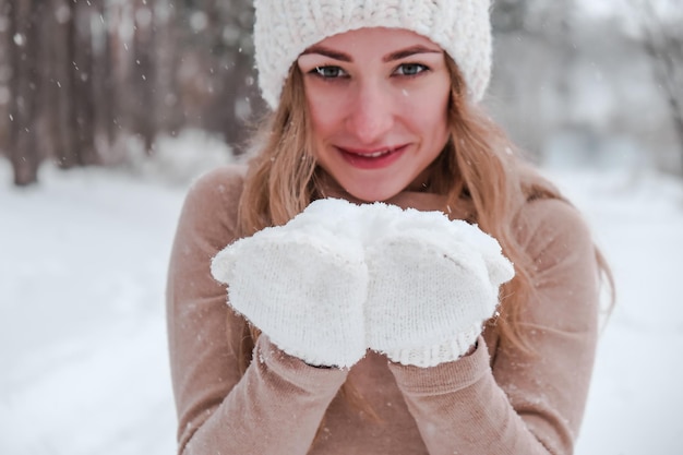 Boże Narodzenie, święta i koncepcja sezonu. Młoda szczęśliwa kobieta wiejący śnieg w zimowym lesie natura. Ciepłe rękawiczki i czapka z dzianiny. Zimowy las krajobraz tło
