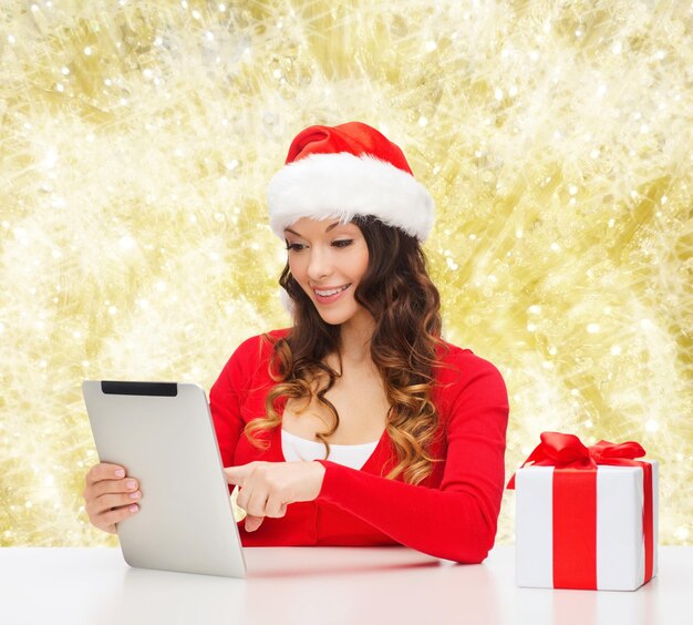 Boże Narodzenie, święta, technologia i koncepcja ludzi - uśmiechnięta kobieta w kapeluszu pomocnika świętego mikołaja z pudełkiem prezentów i komputerem typu tablet na niebieskim tle światła