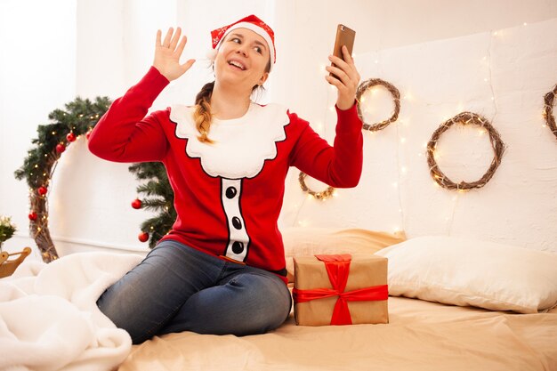 Boże Narodzenie w Internecie Gratulacje z okazji wakacji. Kobieta w radosnym kapeluszu trzyma telefon i komunikuje się z przyjaciółmi i rodziną za pomocą połączenia wideo