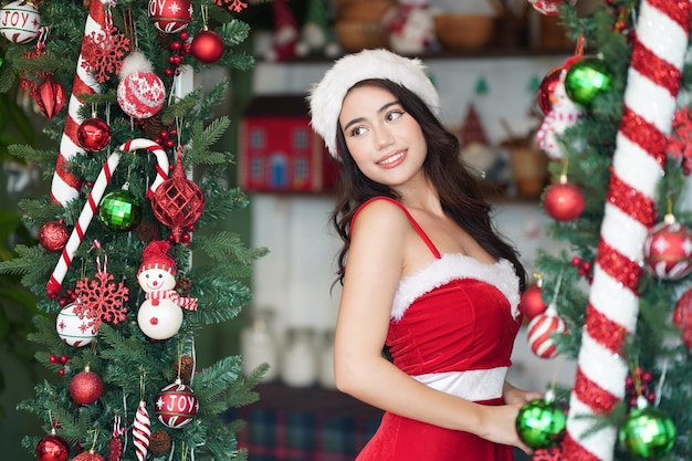 Boże Narodzenie zrelaksowana elegancka młoda kobieta z mikołajowym zwyczajem w czerwonej sukience Boże Narodzenie zrelaksowana elegancka młoda kobieta z mikołajowym zwyczajem w czerwonej sukience i choinką w nowoczesnym domu