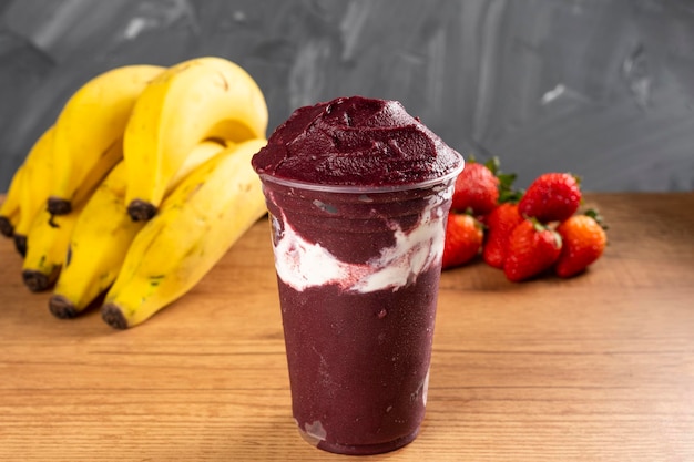 Brazilian Frozen Açai Berry Ice Cream Smoothie w plastikowym kubku ze skondensowanym mlekiem. Na drewnianym biurku i szarym letnim tle z owocami. Widok z przodu dla menu i mediów społecznościowych