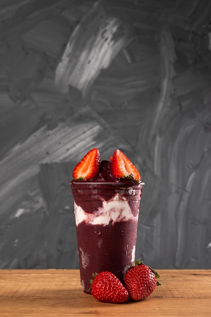 Brazilian Frozen Açai Berry Ice Cream Smoothie w plastikowym kubku ze słomkowymi jagodami i skondensowanym mlekiem. Na drewnianym biurku i szarym letnim tle. Widok z przodu dla menu i mediów społecznościowych